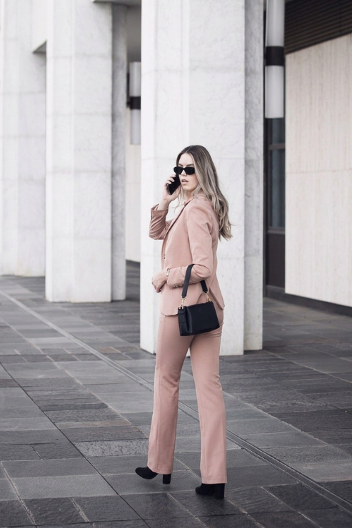 A woman wearing a matching blush pink pants suit and black sunglasses stands on a paved walkway in front of a modern building with large white columns. She is holding a phone to her ear and carrying a small black handbag.