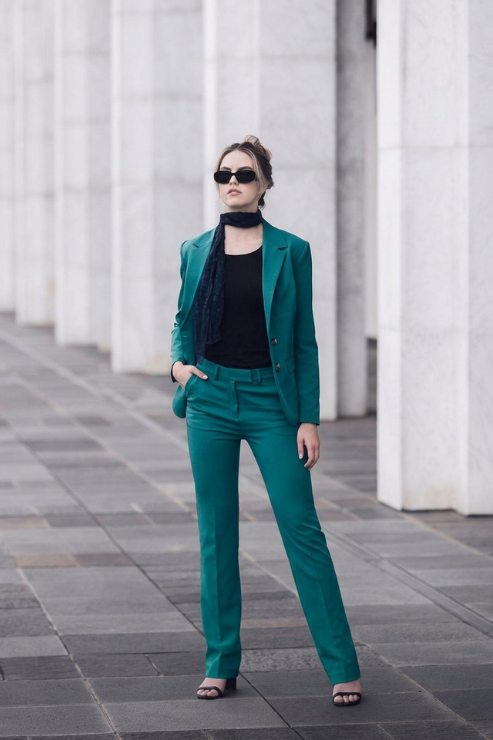 A woman wearing a matching emerald green tailored pantsuit with a black top and scarf, standing outdoors in front of a modern building with white columns.
