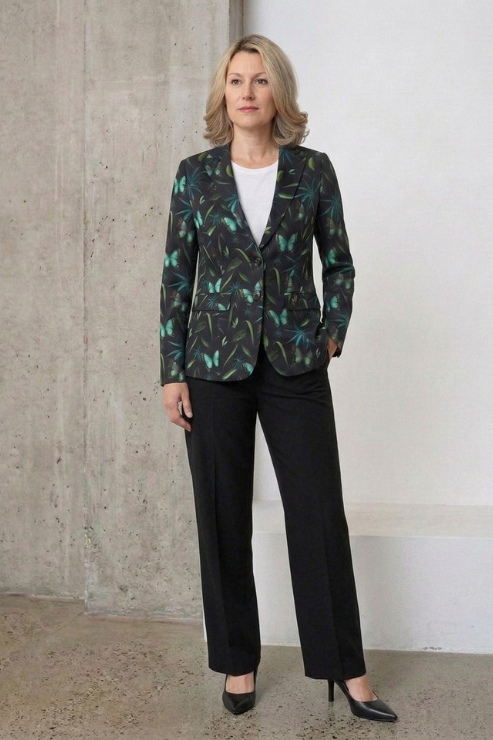 A woman stands indoors wearing a dark patterned blazer with teal and green leaf motifs, black wide-leg trousers, a white top, and black pointed heels against a concrete and white wall background.