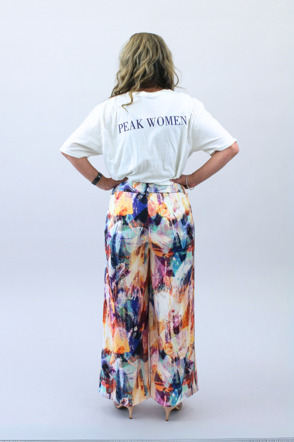 Person wearing a white t-shirt with 'PEAK WOMEN' text and colorful pants on a plain background