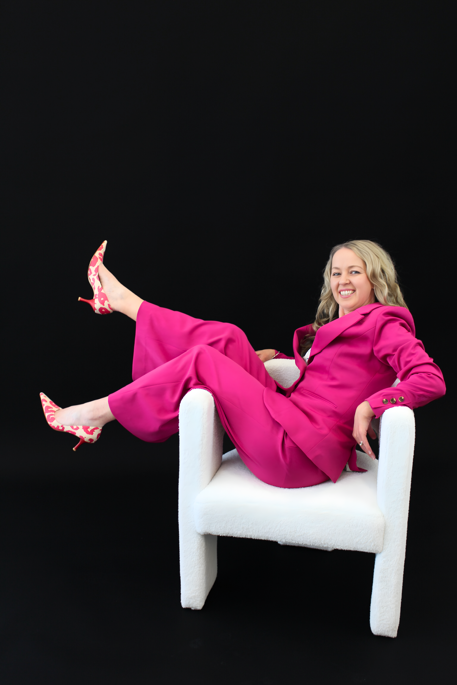 Woman in a pink suit sitting on a white chair against a black background
