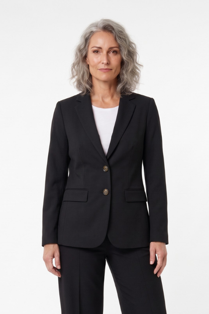 A woman with gray hair wearing a black tailored blazer with two gold buttons over a white top and black trousers, standing against a plain white studio background.
