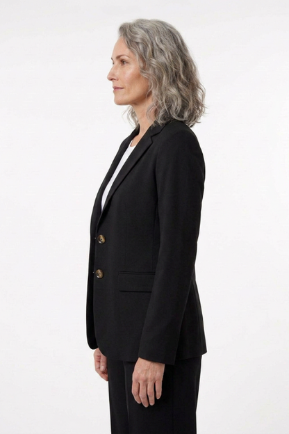 A woman with gray hair is shown in profile wearing a black double-breasted blazer with gold buttons over a white top and black trousers, photographed against a plain white studio background.