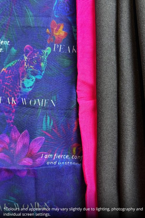 A close-up of a scarf or fabric accessory showing a colorful printed side with a blue-purple background, floral and animal motifs, and white text, next to a bright hot pink fold and a dark gray textured fabric fold.