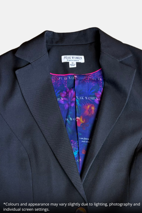 A close-up of a black blazer jacket with a colorful printed inner neck lining visible at the collar, laid flat on a speckled stone surface. The neck label reads 'PEAK WOMEN'.