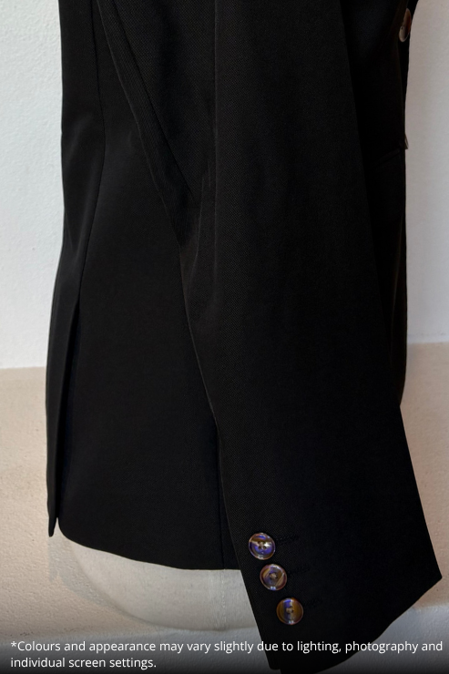 Close-up of a black tailored jacket or blazer sleeve on a mannequin, showing the fabric texture, a vertical seam, and three decorative buttons near the cuff. A small text note at the bottom explains color variations due to lighting.