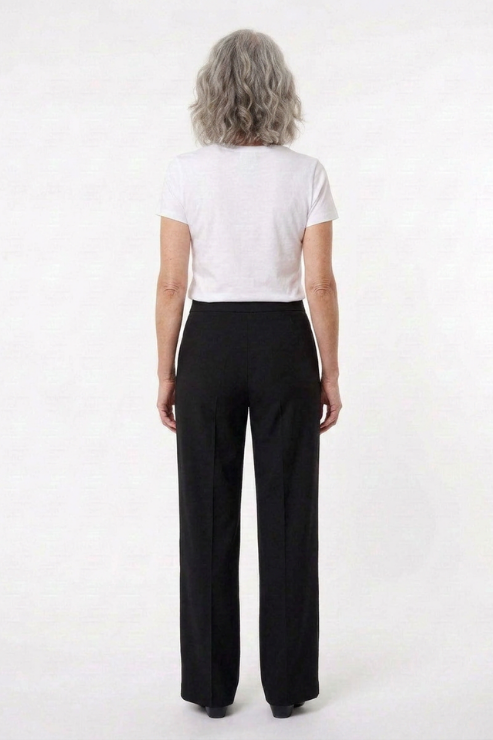 A woman with shoulder-length gray hair is shown from the back, wearing a white short-sleeve crop top and black straight-leg trousers against a plain white studio background.