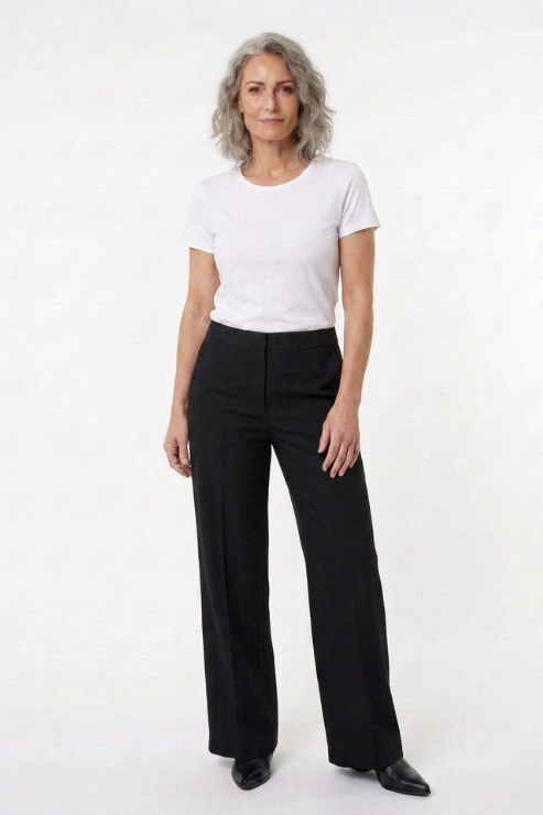 A woman with gray hair stands against a plain white studio background wearing a white short-sleeve crewneck t-shirt and black wide-leg trousers.