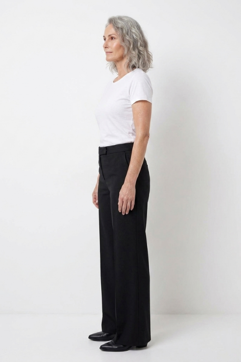 A woman with gray hair stands in profile wearing a white short-sleeve t-shirt and black straight-leg trousers against a plain white studio background.