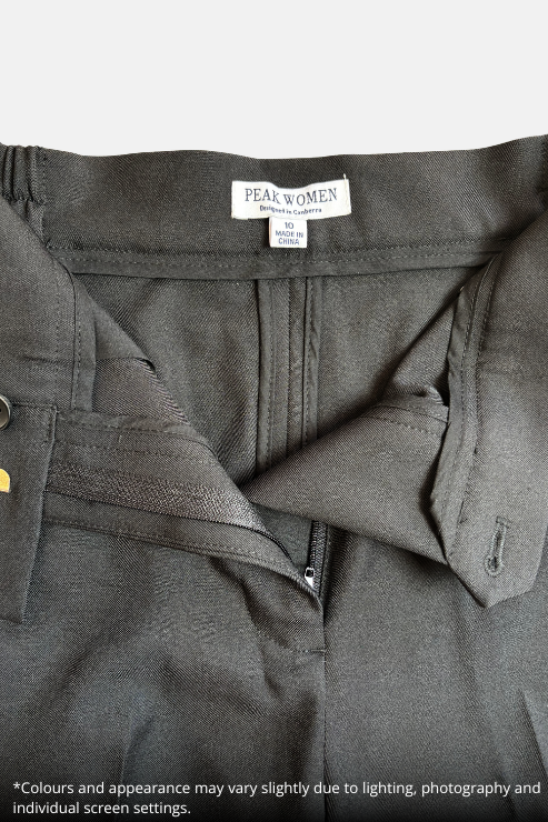 Close-up of a black zippered work jacket or uniform top by Peak Women, showing the inner collar label and zipper detail, laid flat on a speckled beige countertop.