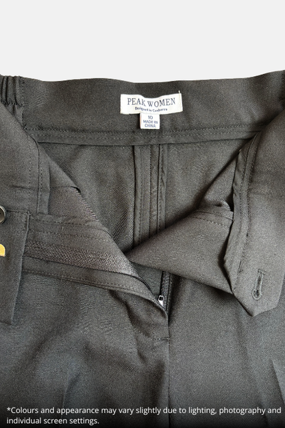 Close-up of a black zippered work jacket or uniform top by Peak Women, showing the inner collar label and zipper detail, laid flat on a speckled beige countertop.