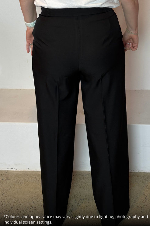 Black wide-leg trousers shown on a model from the back, featuring a high waistband and front pleats. The model is wearing a white top, and the photo is taken against a plain indoor wall with a beige floor.