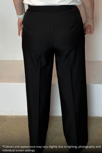 Black wide-leg trousers shown on a model from the back, featuring a high waistband and front pleats. The model is wearing a white top, and the photo is taken against a plain indoor wall with a beige floor.