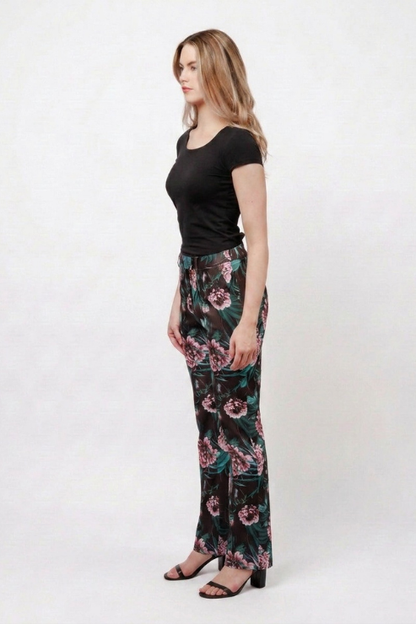 A woman stands in profile wearing a black short-sleeve top and high-waisted, wide-leg trousers with a dark floral print in pink, green, and black. She is wearing black heeled sandals against a plain white studio background.