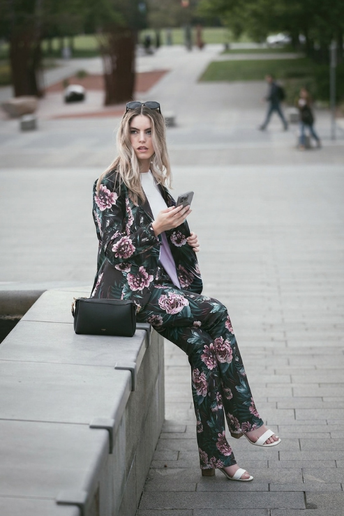 A woman sits on a concrete ledge in an outdoor urban setting, wearing a matching black floral blazer and wide-leg trousers set with pink and purple flowers. She is holding a phone and has a black handbag resting beside her.