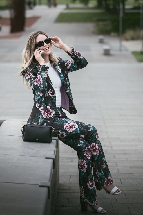 A woman wearing a dark green floral blazer and matching wide-leg pants sits on a concrete bench in an outdoor park setting. She is wearing a white top, black sunglasses, and white heeled sandals, with a black handbag resting beside her.