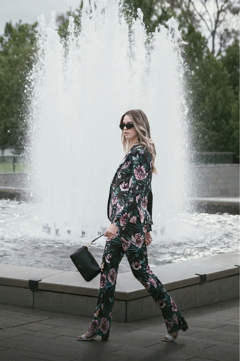 A woman walks in front of a large fountain wearing a dark floral jumpsuit with pink and light blue flowers, black sunglasses, and carrying a black handbag.