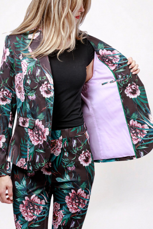 A woman models a matching floral blazer and trousers set. The fabric has a dark brown base with a vibrant pink and green tropical floral print.