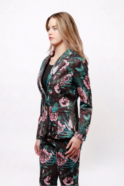 A woman modeling a matching floral blazer and trousers set. The suit features a dark background with a vibrant pink and green tropical floral print, styled over a black top against a plain white studio background.