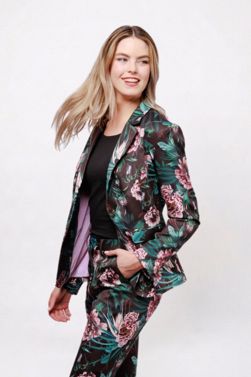 A woman modeling a matching floral blazer and trousers suit set. The suit features a dark green base with pink and purple floral patterns, worn over a black top and a light purple shirt.