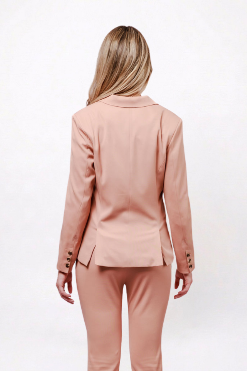 A woman viewed from the back wearing a matching blush pink tailored blazer and trousers set against a plain white studio background.