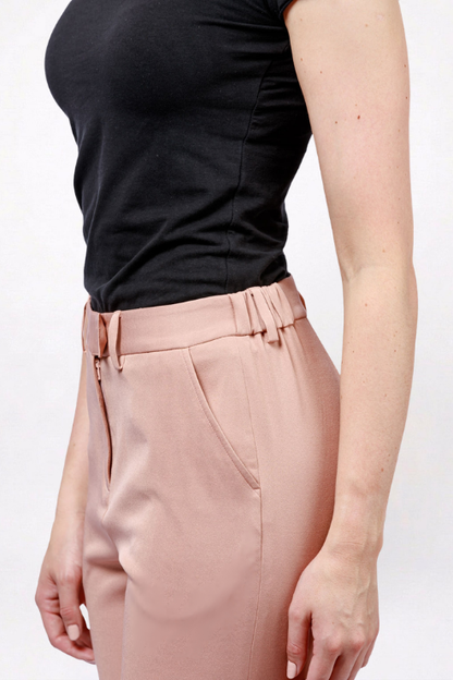A close-up side view of a person wearing high-waisted blush pink trousers with a matching belt and pleated front detail, paired with a black sleeveless top against a plain light background.