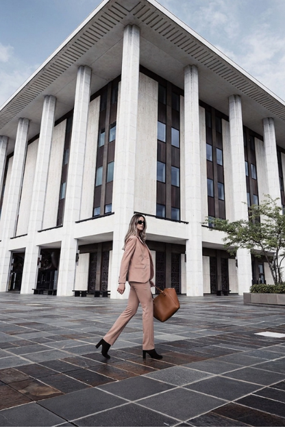 A woman in a beige two-piece suit walks across a paved plaza in front of a large modern building with white columns and dark window panels, carrying a brown leather tote bag.