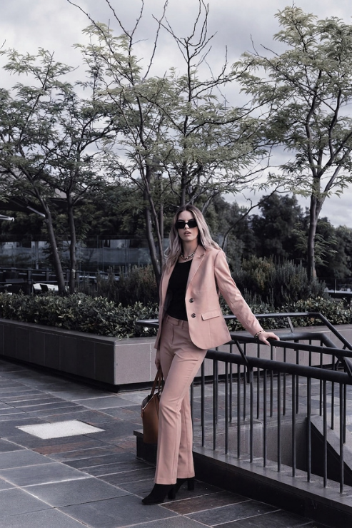 A woman stands outdoors on a paved walkway beside a black metal railing, wearing a light blush pink tailored blazer and matching trousers over a black top, with black ankle boots and a brown handbag.