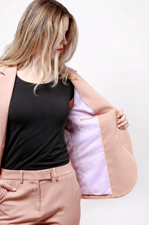 A woman wearing a peach-colored blazer over a black top and matching peach trousers, holding the blazer open to show the light pink lining.