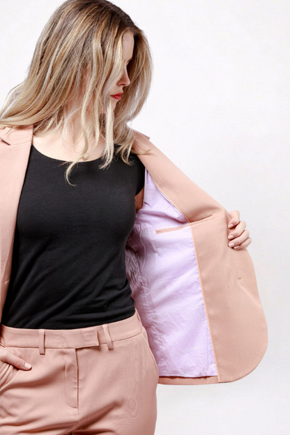 A woman wearing a peach-colored blazer over a black top and matching peach trousers, holding the blazer open to show the light pink lining.
