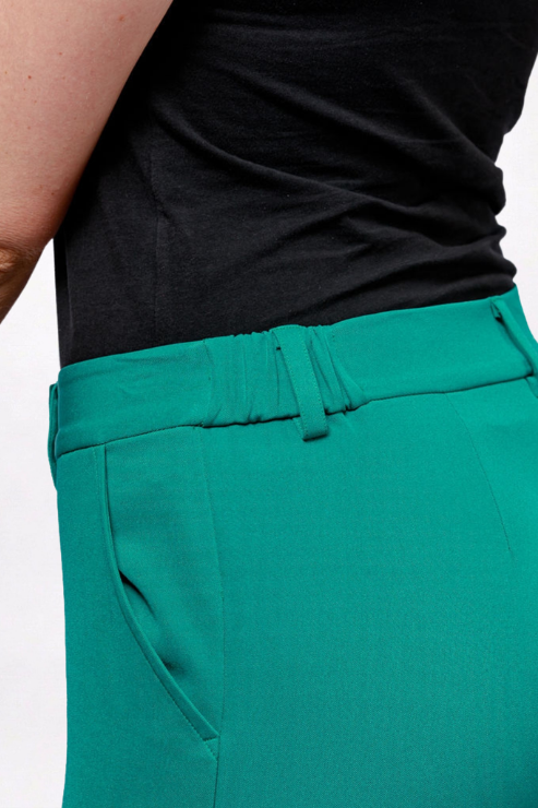 Close-up of a person wearing bright teal-green tailored trousers and a black top, cropped to the waist area against a plain light background.