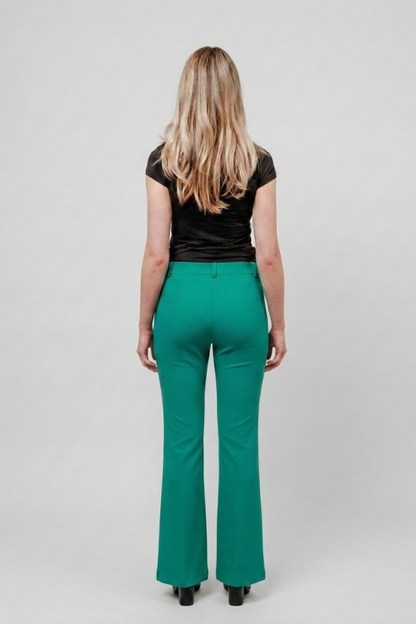 A woman viewed from the back wearing bright green flared pants, a black short-sleeve top, and black heels against a light gray studio background.