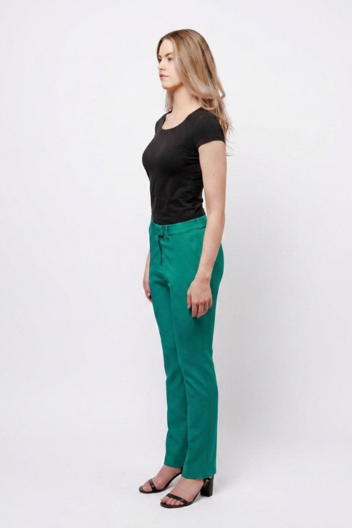 A woman standing in profile wearing a black short-sleeve top, bright green trousers, and black heeled sandals against a plain light gray studio background.