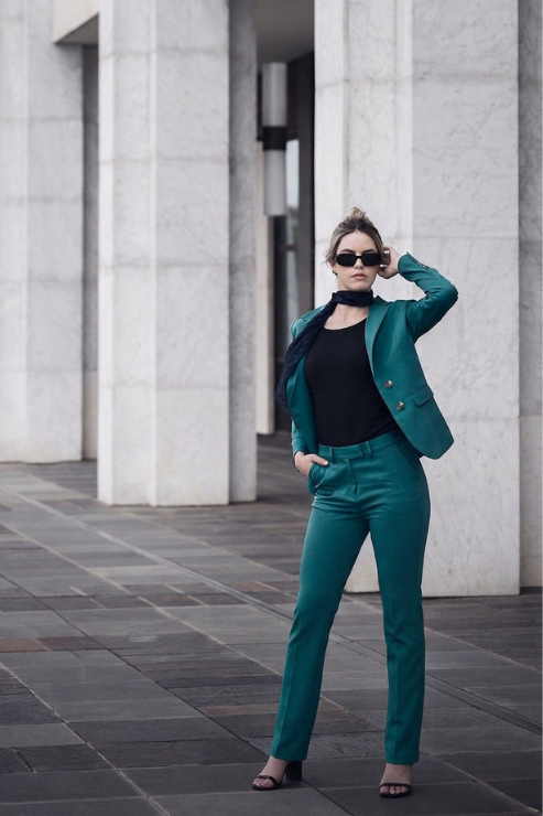 A woman posing in a stylish teal green tailored pantsuit with a black top and sunglasses, standing in front of a modern building with stone columns.