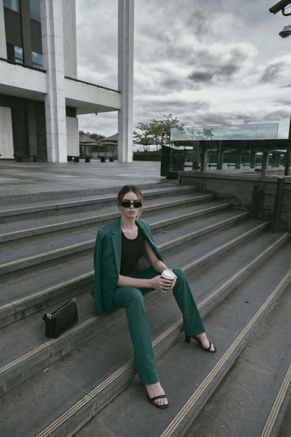 A woman wearing a matching emerald green blazer and trousers set sits on outdoor concrete steps, holding a takeaway coffee cup. She is wearing dark sunglasses and black sandals, with a small black handbag resting beside her.