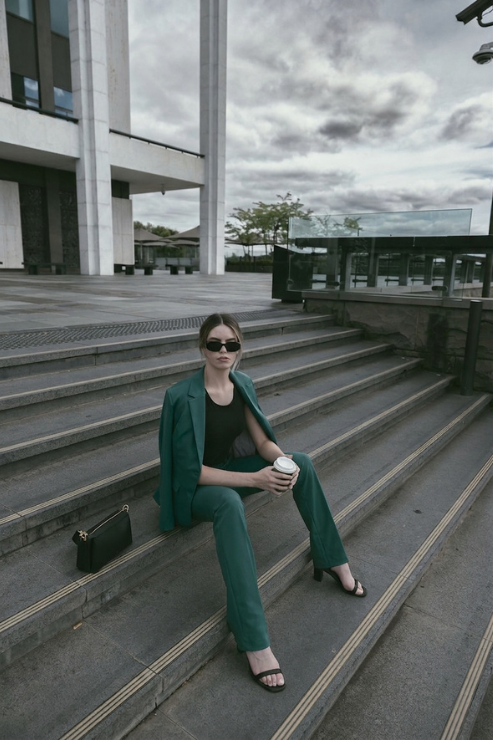 A woman wearing a matching emerald green blazer and trousers set sits on outdoor concrete steps, holding a takeaway coffee cup. She is wearing dark sunglasses and black sandals, with a small black handbag resting beside her.