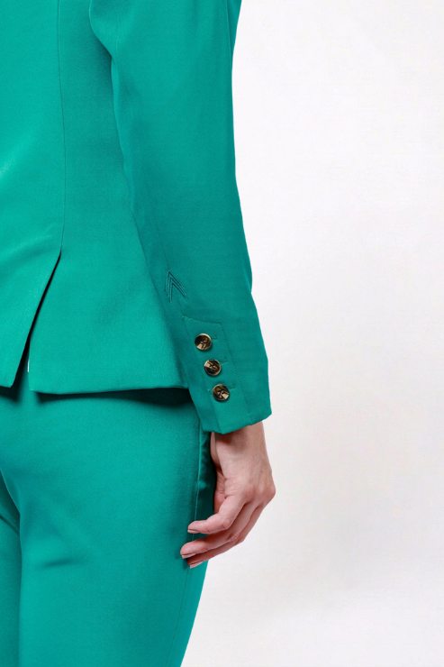Close-up of a person wearing a tailored teal-green suit jacket and matching trousers, showing the sleeve with three gold buttons and the hand holding the hem.