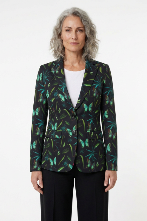 A woman with gray hair stands against a white studio background wearing a dark blazer with a tropical butterfly and palm leaf print in green and teal, layered over a white top and black trousers.