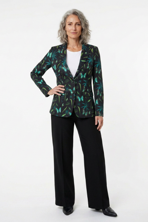 A woman modeling a black blazer with a green and teal tropical leaf pattern, worn over a white top and paired with black wide-leg trousers, against a plain white studio background.