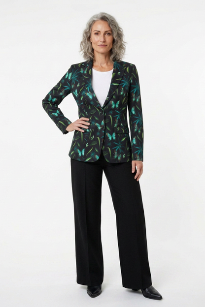A woman modeling a black blazer with a green and teal tropical leaf pattern, worn over a white top and paired with black wide-leg trousers, against a plain white studio background.