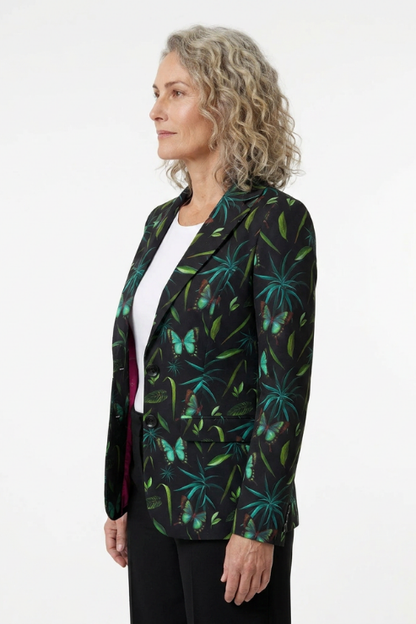 A woman with curly blonde hair is shown in profile wearing a black blazer with a vibrant green and teal tropical leaf and butterfly pattern, layered over a white top and black trousers, against a plain white studio background.