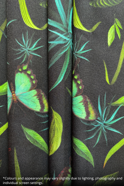 A close-up of a dark charcoal gray fabric with a tropical print featuring green and turquoise butterflies and leaves. The fabric is shown in soft vertical folds, and a small text disclaimer is visible at the bottom left corner.