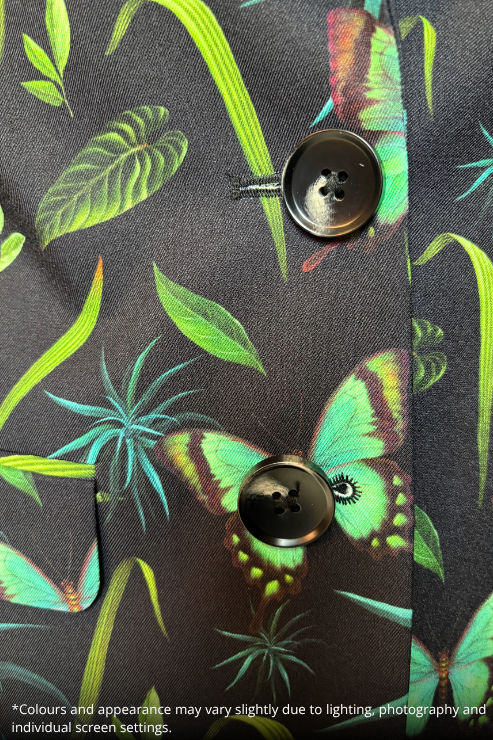 Close-up of a dark charcoal gray button-up shirt featuring a vibrant tropical print with green leaves, turquoise plants, and green-and-black butterflies. The shirt has two dark brown buttons and a visible pocket seam on the left side.
