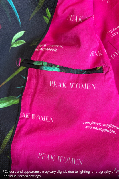 Close-up of a bright pink fabric garment with repeated 'PEAK WOMEN' text and a black zipper detail, laid over a dark green patterned fabric background.