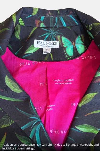 Close-up of a Peak Women Mirabel blazer or garment showing a black tropical leaf and butterfly print outer layer with a bright pink inner lining and a visible Peak Women neck label.